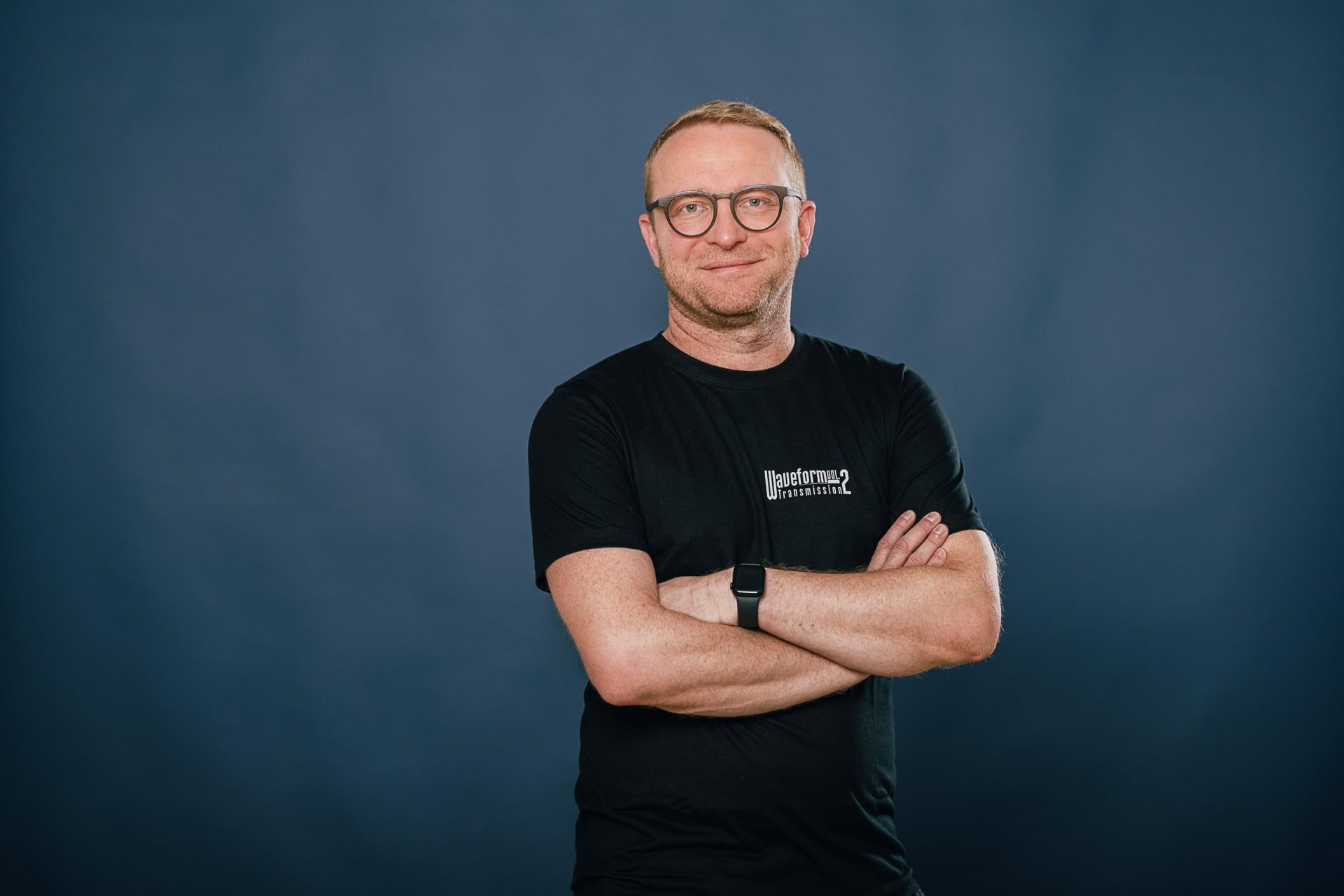 Mann mit kurzen blonden Haaren und Brille, trägt ein schwarzes T-Shirt, lächelt in Kamera