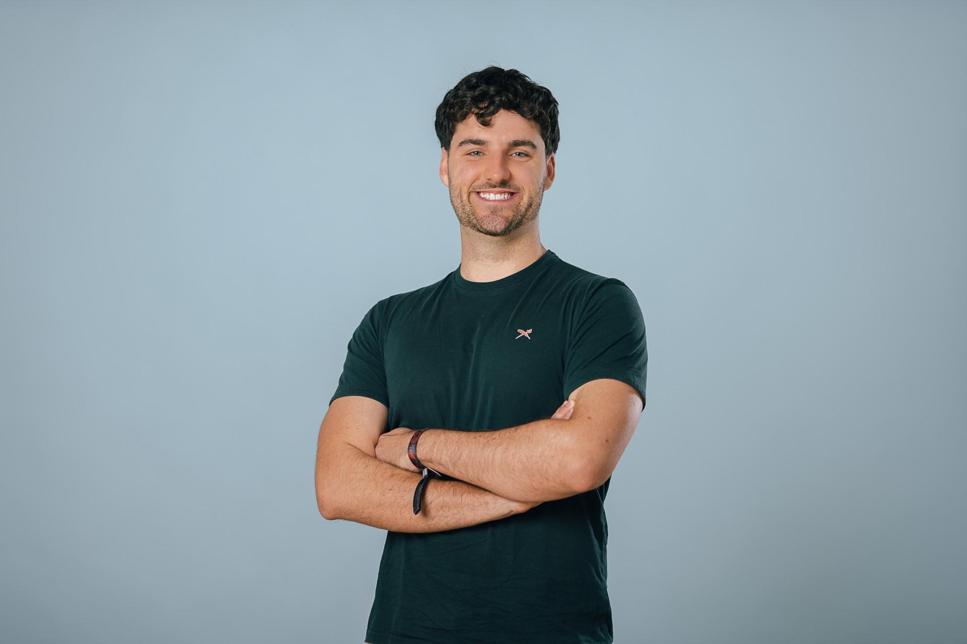 Claudio Bischoff | Werkstudent ZIT Mann mit dunklen gekräuselten Haaren lächelt in Kamera und verschränkt Arme vor T-Shirt